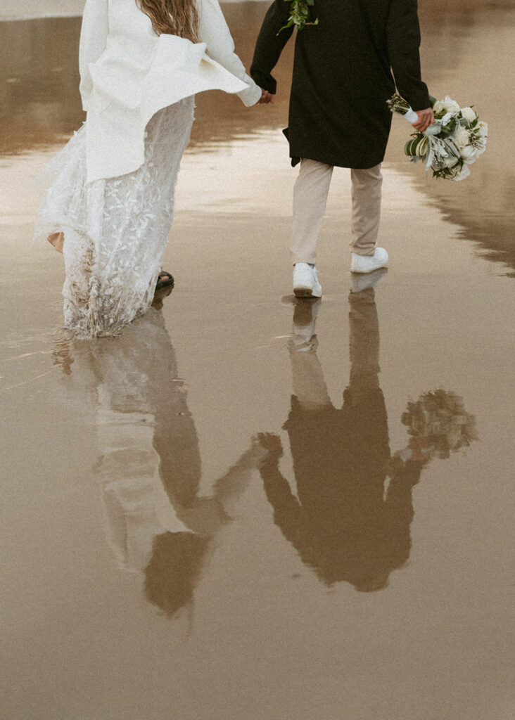 bride and groom holding hands walking along a wet sandy beach