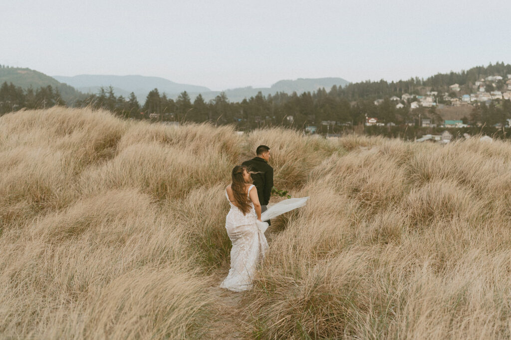 bride and groom in their in wedding attire walks hand-in-hand through tall grass on a sandy path