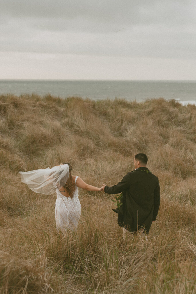 bride in a white dress and veil holding hands with the groom in a dark suit as they walk through tall grass towards a foggy coastline