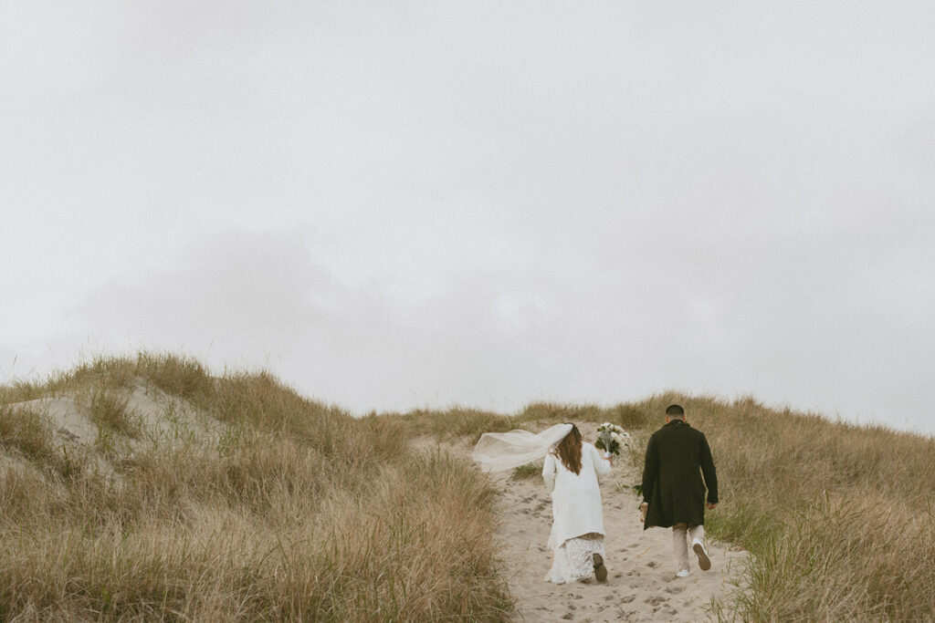 Bride and groom in wedding attire walking in a sandy path between grassy dune