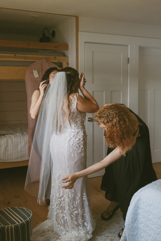 bride preparing in a bedroom wearing a lace wedding dress and veil and other two women are helping