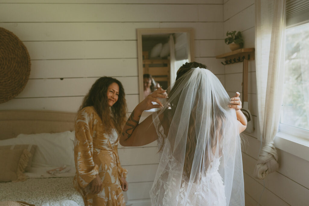 bride wearing a wedding dress and veil stands in front of a mirror adjusting her gown and her mother standing in front