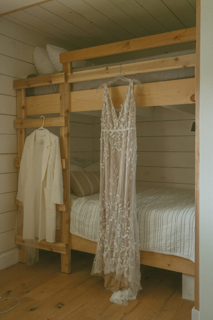 lacey wedding dress hangs from the top bunk of a wooden bunk bed in a cozy, rustic room with shiplap walls