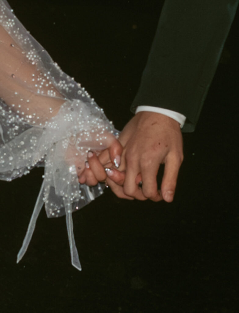 bride wearing a white, pearl-adorned sleeve with delicate ribbons, while the groom in a dark suit and both hands are interlocked