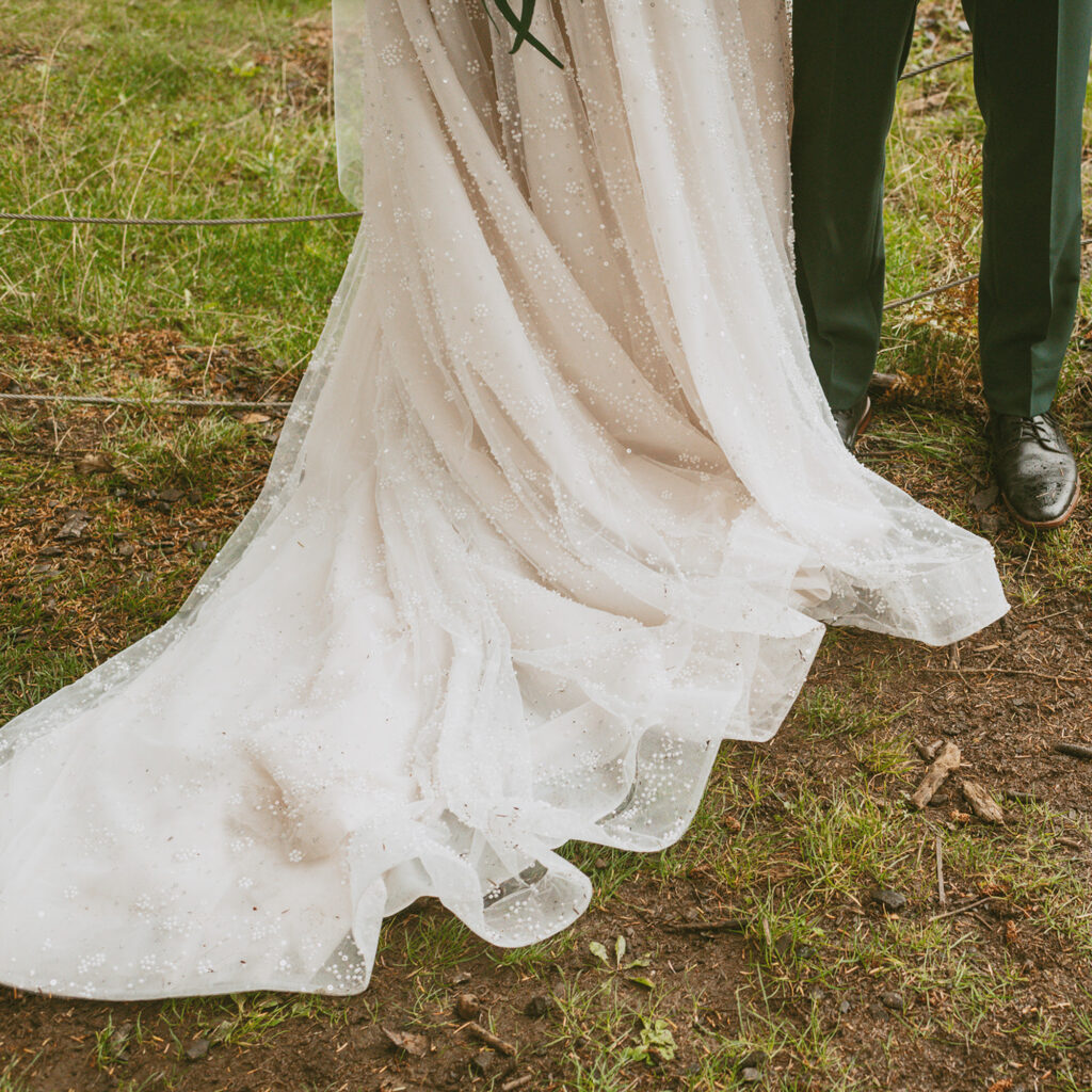 close-up of a bride and groom standing outdoors on grass showing only the lower half of the bride's dress and the groom's green pants and brown shoes