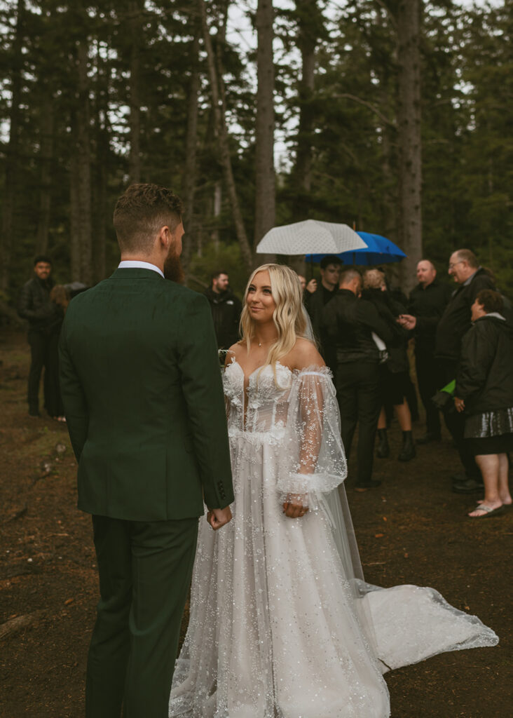 bride and groom facing each other on their outdoor wedding ceremony in a forest while guests holding umbrellas