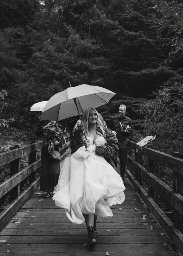 bride wearing her wedding dress holding an umbrella walks happily across a wooden footbridge in a forest