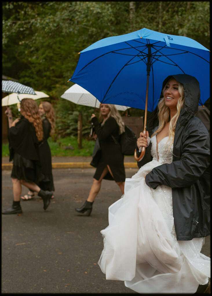 bride in a white dress and black jacket walking outdoor under a blue umbrella with other group of people holding their umbrellas too