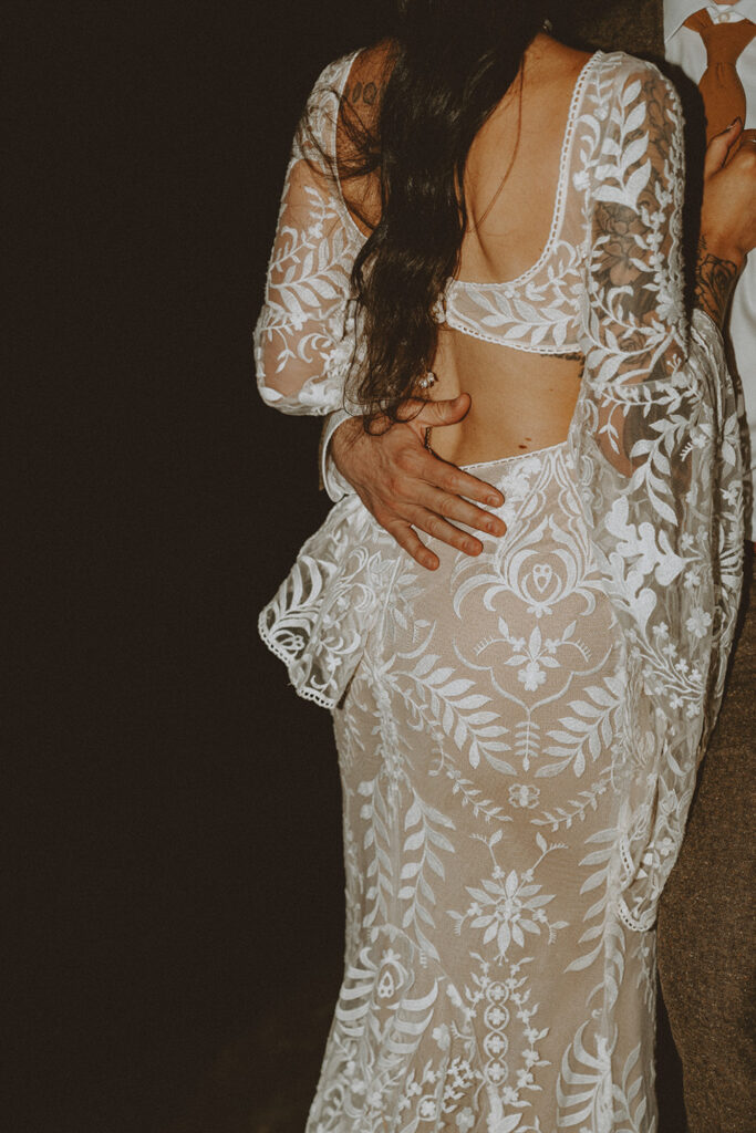 bride in a lace-patterned, backless wedding dress with long sleeves is embracing and being held by someone