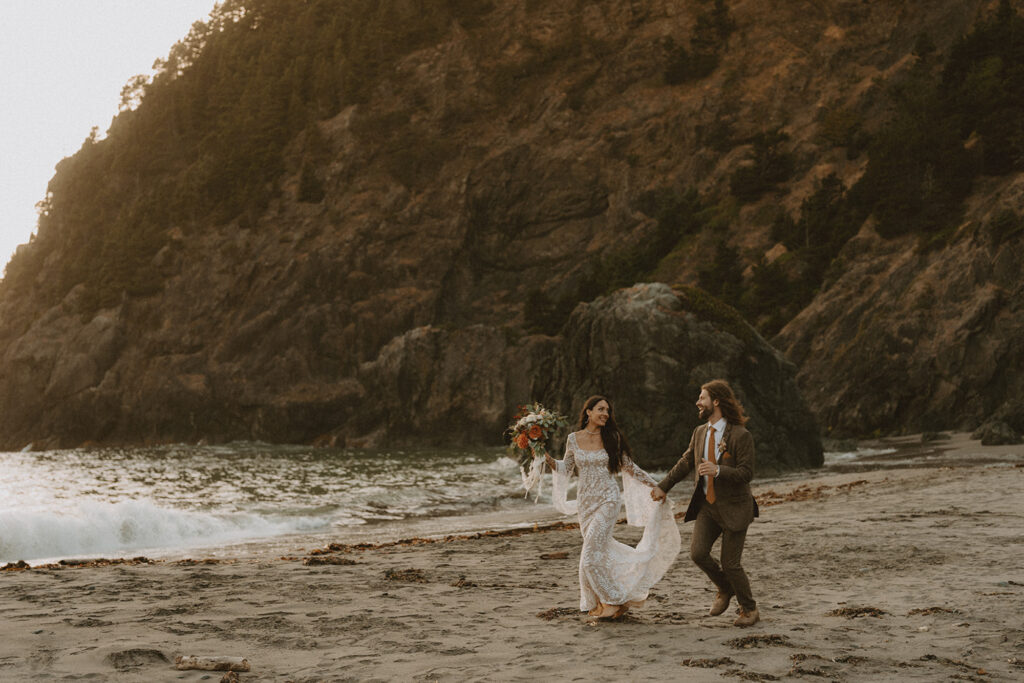 bride in white lacey dress and holding bouquet running alongside her husband in a brown suit on the Oregon coast