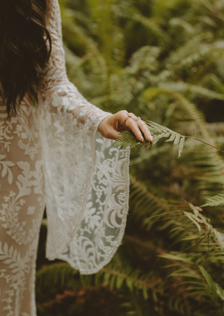 bride wearing a white lace dress with floral patterns holding a fern leaf in a lush, green forest