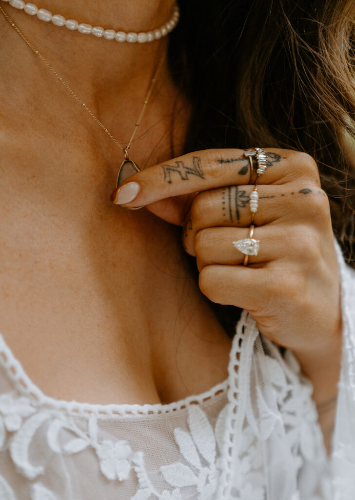 bride wearing her white lace top and a pearl choker holding her necklace with a small pendant and hand has multiple rings and tattoos