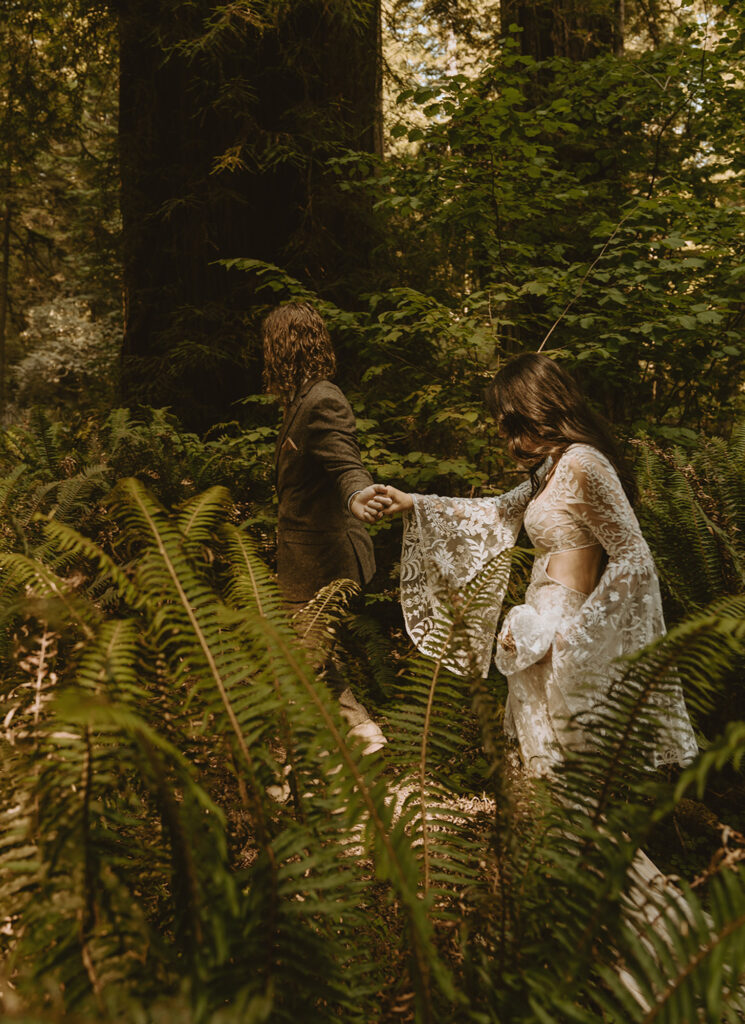 bride and groom walks hand-in-hand through a lush, fern-filled forest