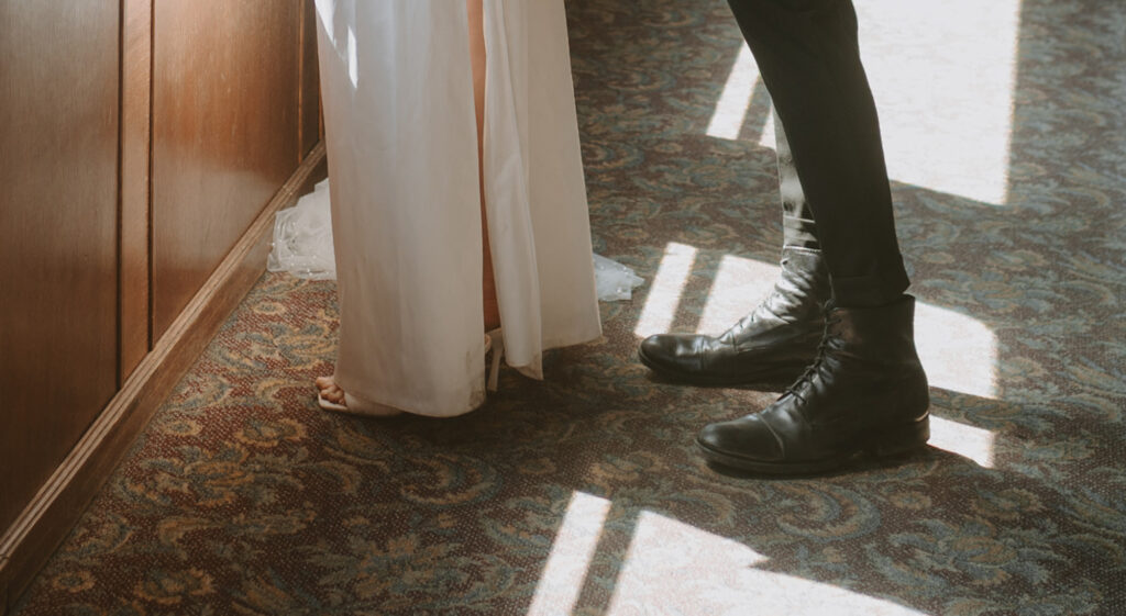close-up photo showing the lower portion of a bride in a white dress, with her feet visible and groom in dark pants and black dress shoes standing together