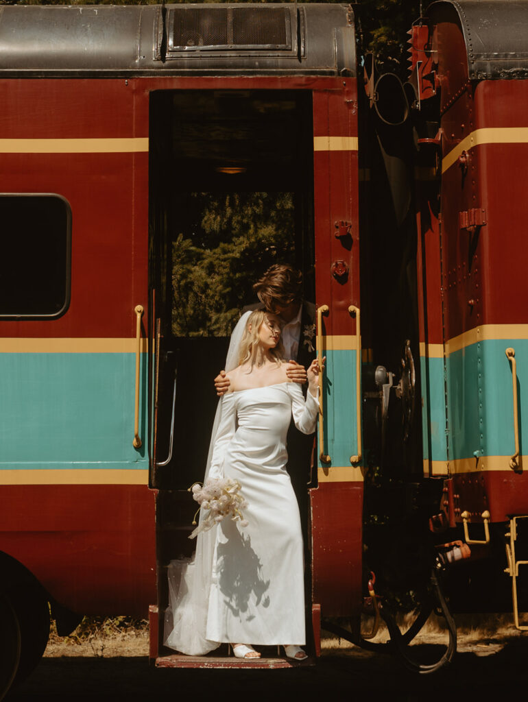 bride in a wedding gown and holding a bouquet stands on the steps of a vintage train, gazing down and groom embraces her while looking over her shoulder