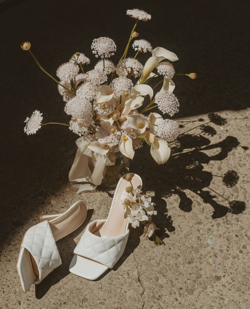 bouquet of white flowers rests on a concrete floor and a pair of white open-toe, quilted slip-on shoes decorated with small white flowers
