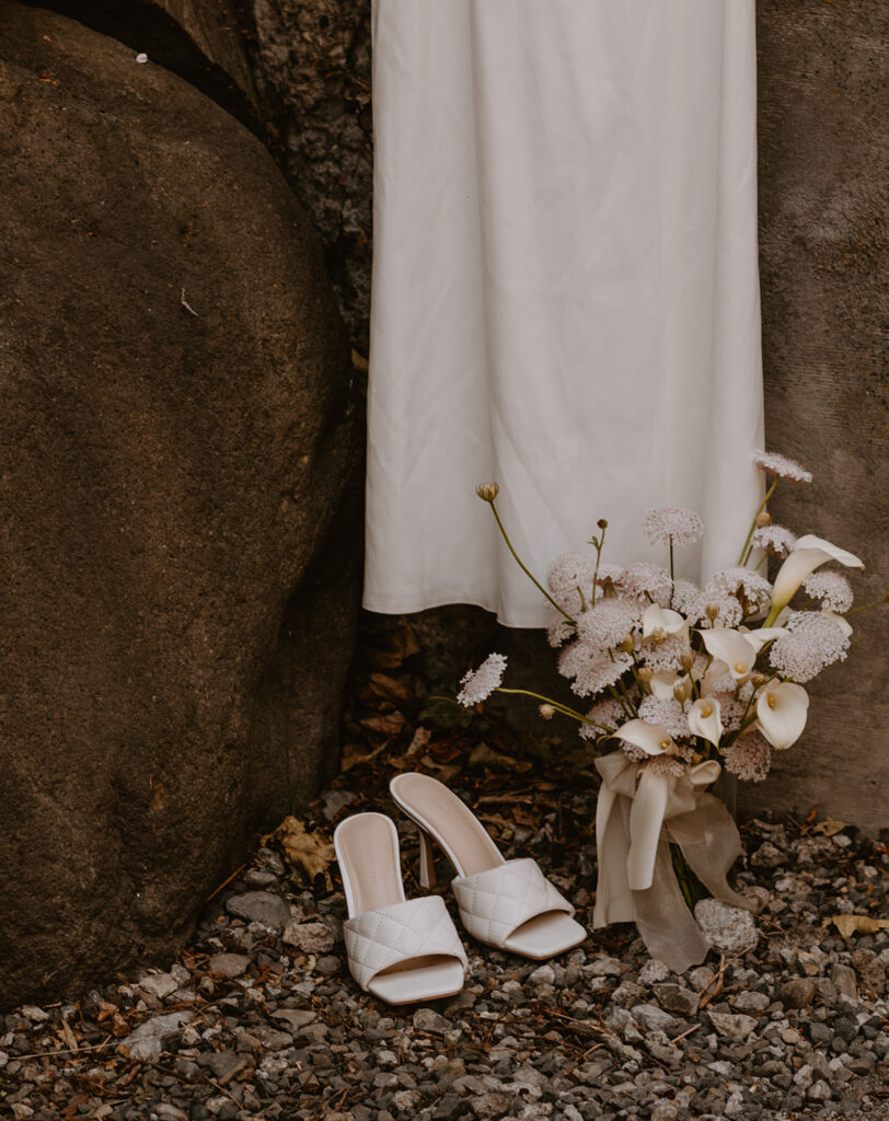 white wedding dress drapes over large rocks, and a pair of white heeled sandals placed on gravel in front