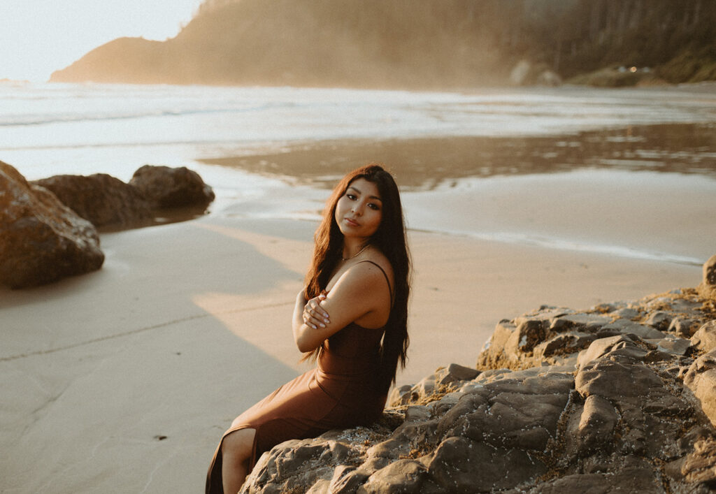 Woman in a long, brown dress sits on rocky terrain at the beach during sunset for her senior beach photo