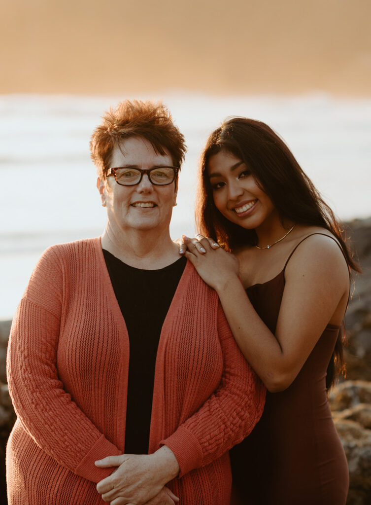 Older woman with short hair and glasses, wearing a red cardigan, stands alongside a younger woman with long hair wearing a sleeveless brown dress, both smiling with a serene beach in the background