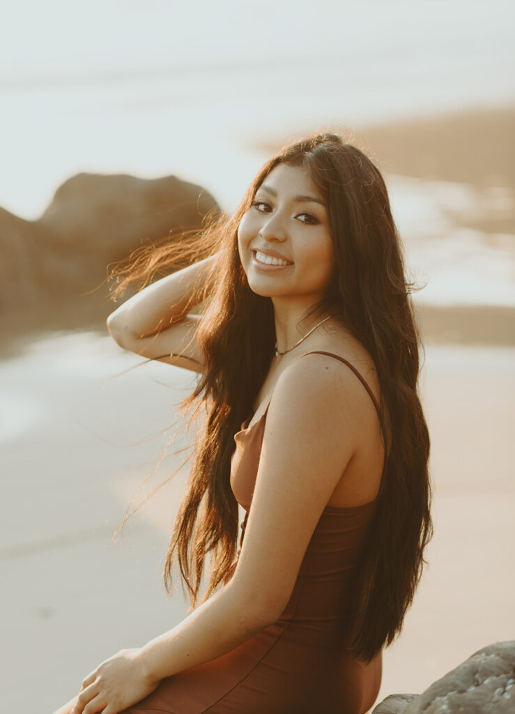 Woman with long dark hair smiles while sitting on the beach at sunset for her senior beach photo
