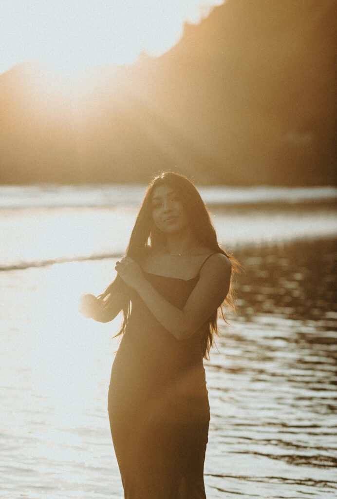 Woman with long hair standing by the water during sunset, dressed in a dark, sleeveless dress, smiling slightly