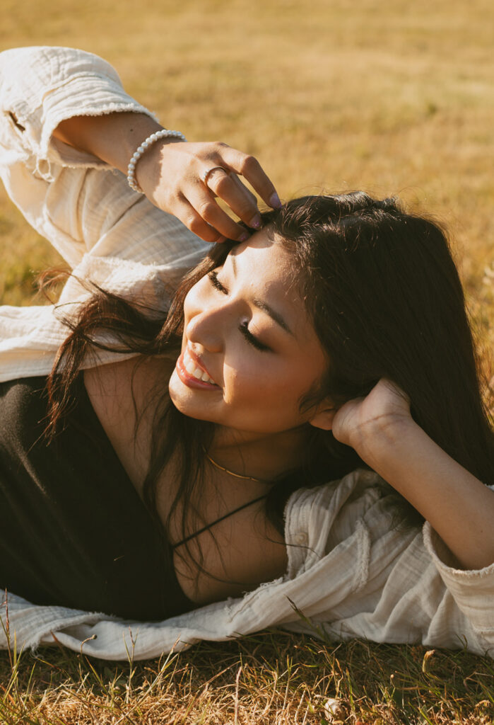 Woman with long dark hair lying on a grassy field smiling while holding a strand of her hair in one hand and resting her other hand behind her head