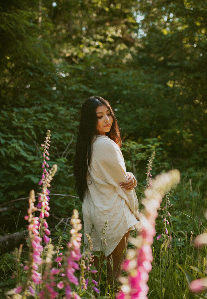 Woman with long dark hair stands in a sunlit forest clearing, gazes down with a gentle expression, surrounded by tall pink wildflowers and lush greenery