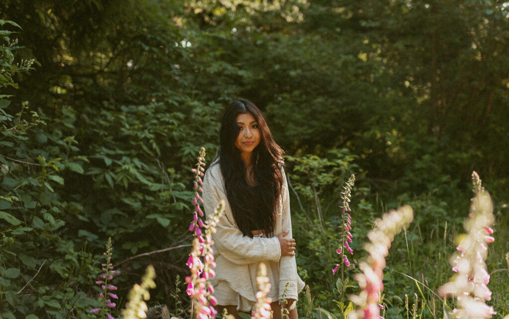 Woman with long dark hair stands in a sunlit forest clearing, surrounded by tall pink wildflowers and lush greenery