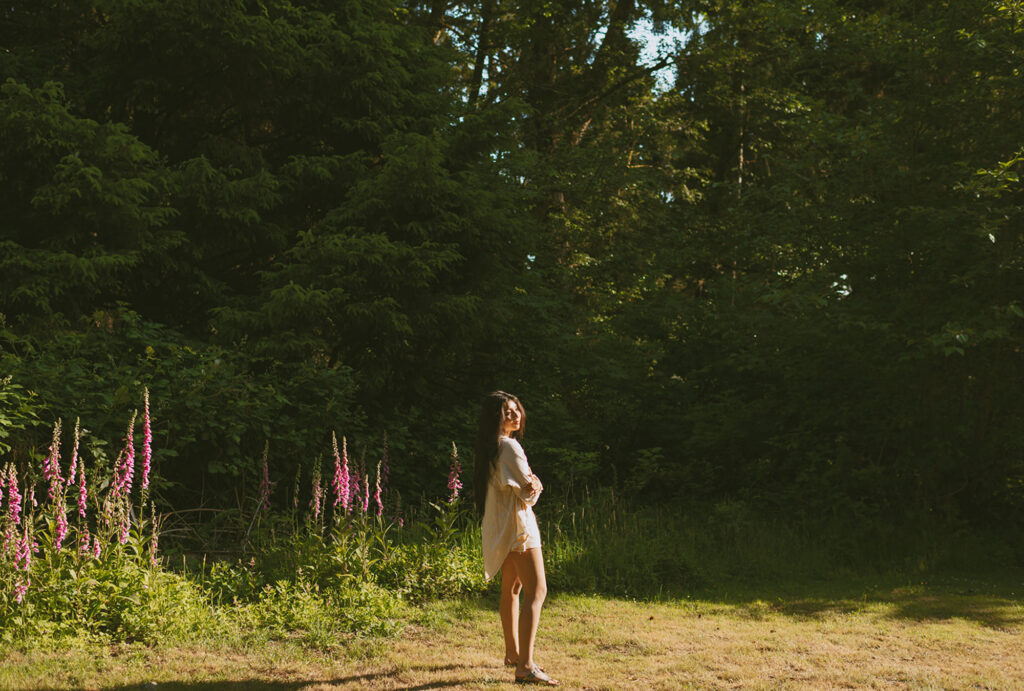 Woman standing in a sunlit clearing surrounded by tall trees and pink flowers 