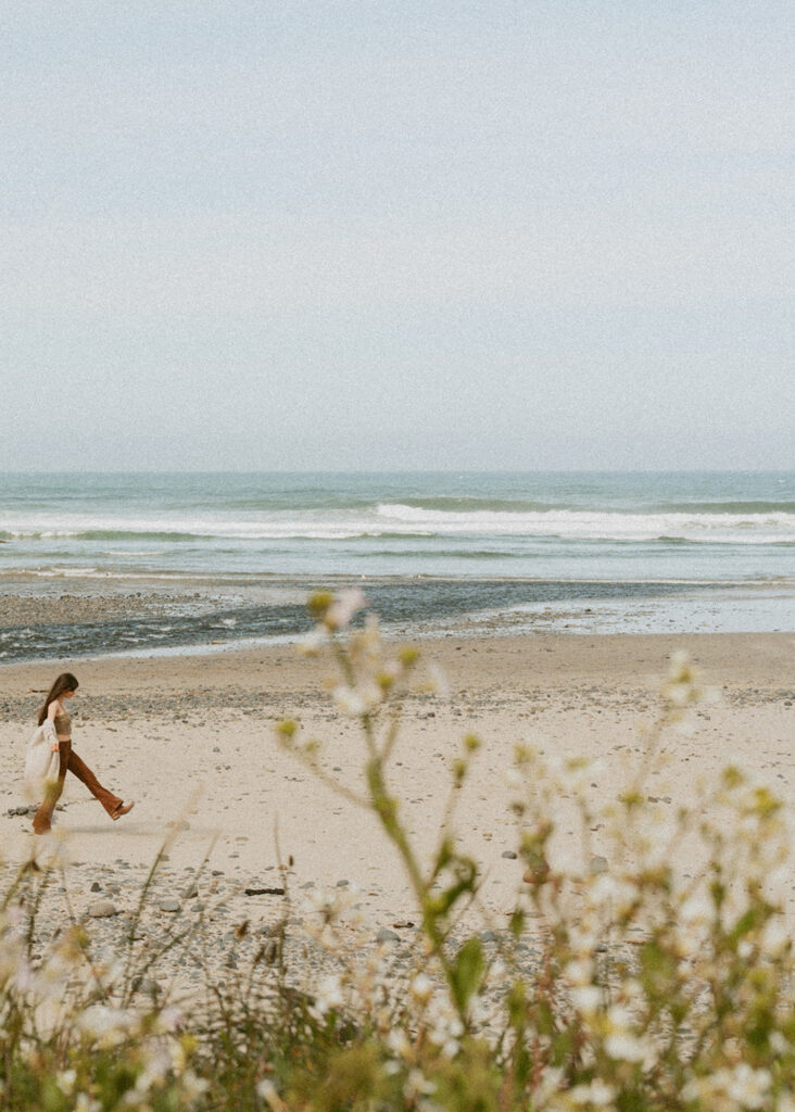 Woman walking along a sandy beach with gentle waves in the background in Florence, Oregon for her senior beach photo shoot