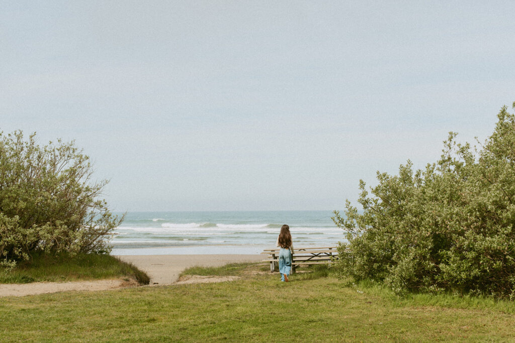 Woman standing at the edge of a grassy area, looking out at the ocean waves