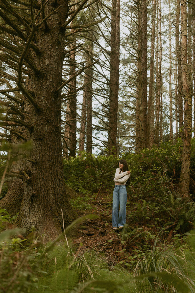 Woman standing alone in a forest filled with tall trees and greenery, wearing a sweater and jeans