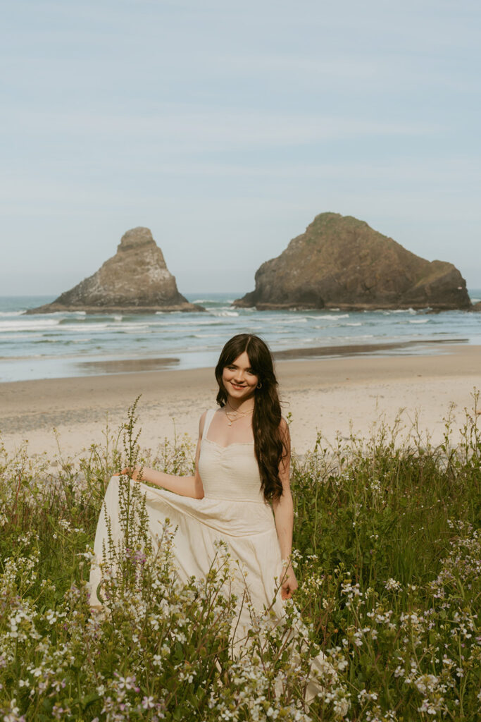 Woman with long dark hair and a white dress stands in a field of wildflowers on a beach