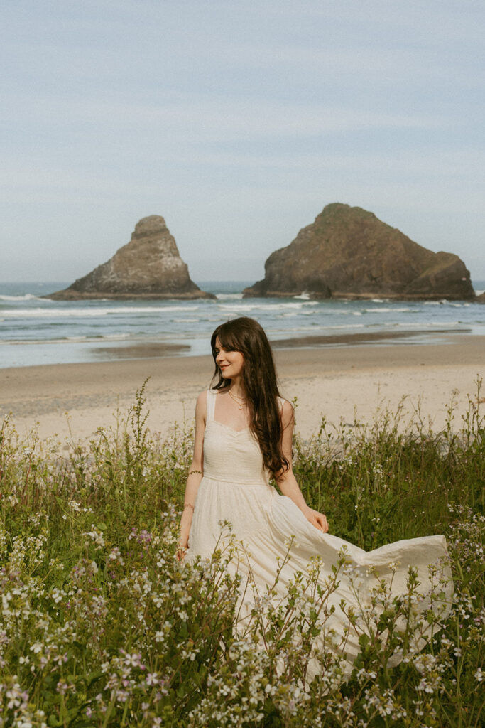 Woman in a flowing white dress standing in a field of wildflowers on a beach for her senior beach photo shoot