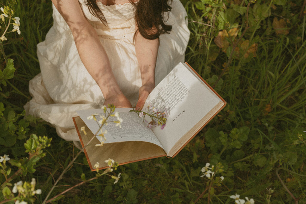Woman in a white dress sits on grass in a lush garden, holding an open book