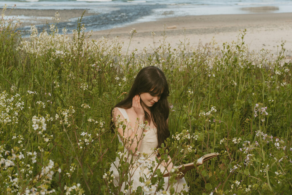 Woman with long dark hair dressed in a light-colored top is sitting amongst tall wildflowers on a grassy dune near the beach, reading a book