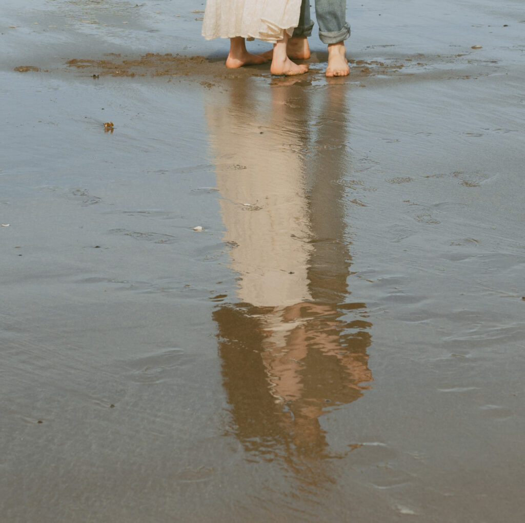 Partial view of two people walking barefoot on the beach, reflecting on the wet sand