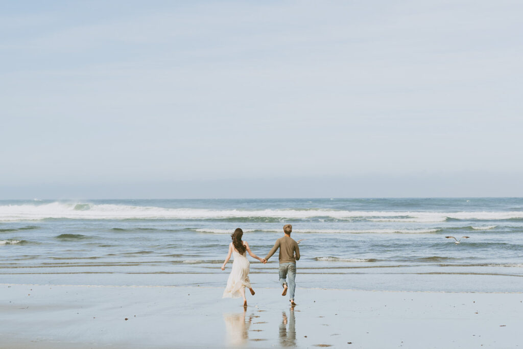 A couple holding hands and running towards the ocean on a sandy beach in Florence, Oregon