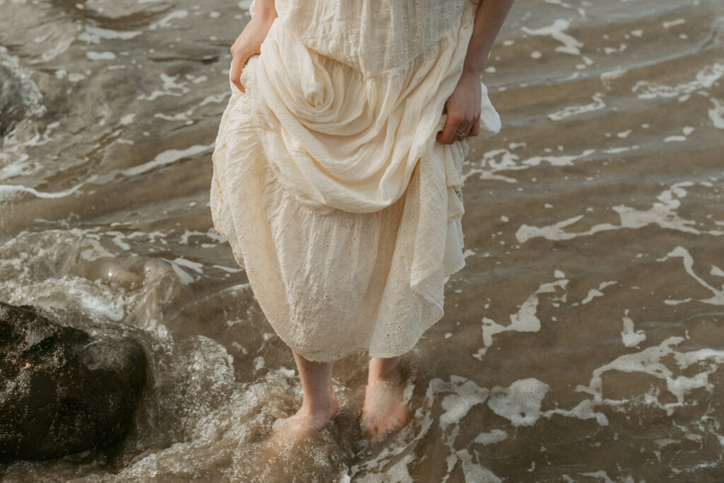 Woman wearing a flowing white dress stands barefoot on a sandy beach, with gentle waves lapping around their feet