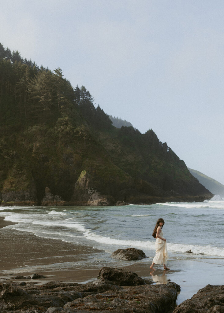A woman in a flowing white dress walking along a rocky shoreline with gentle waves lapping at her feet