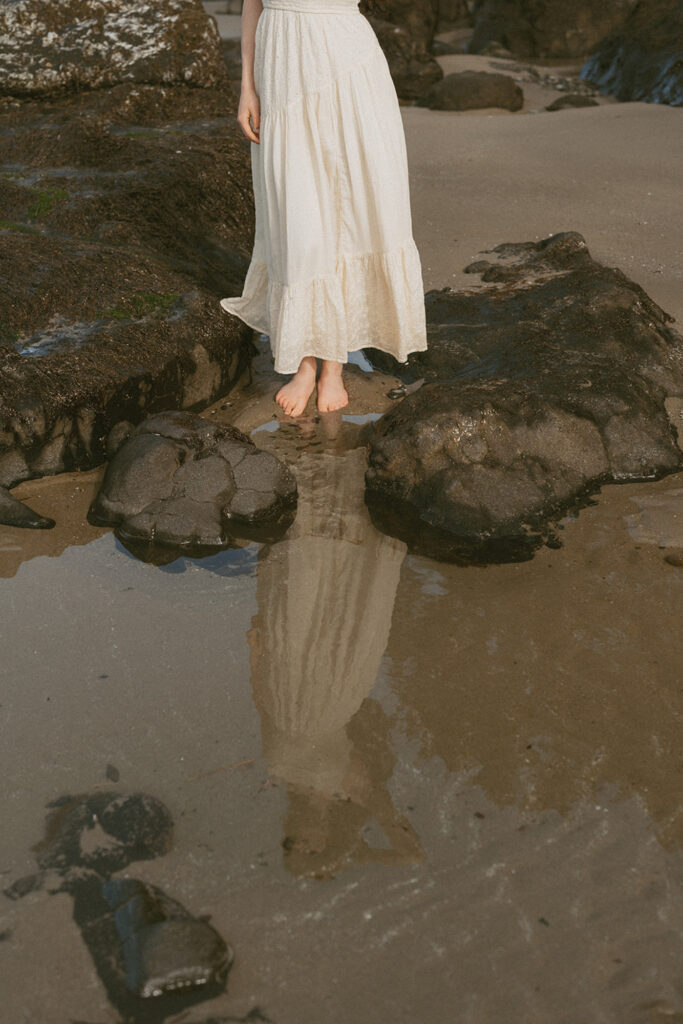 Woman in a long, flowing white skirt standing barefoot on wet sand between dark rocks at the beach in Florence, Oregon