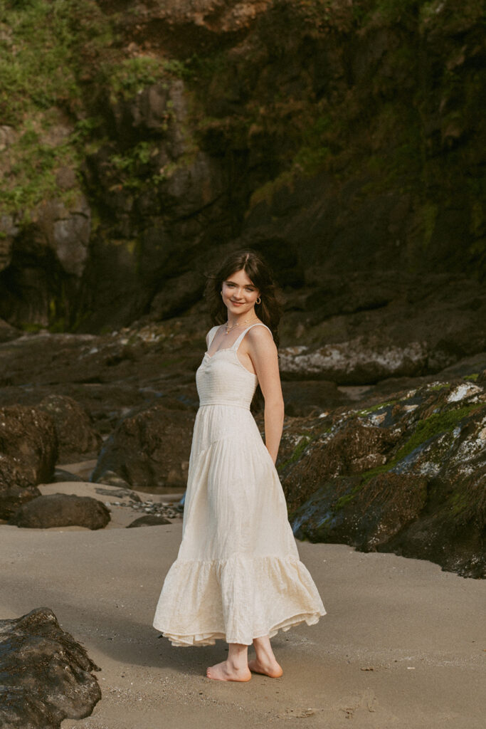 Woman in a long, white dress strolls barefoot on a beach, with rocks and greenery in the background