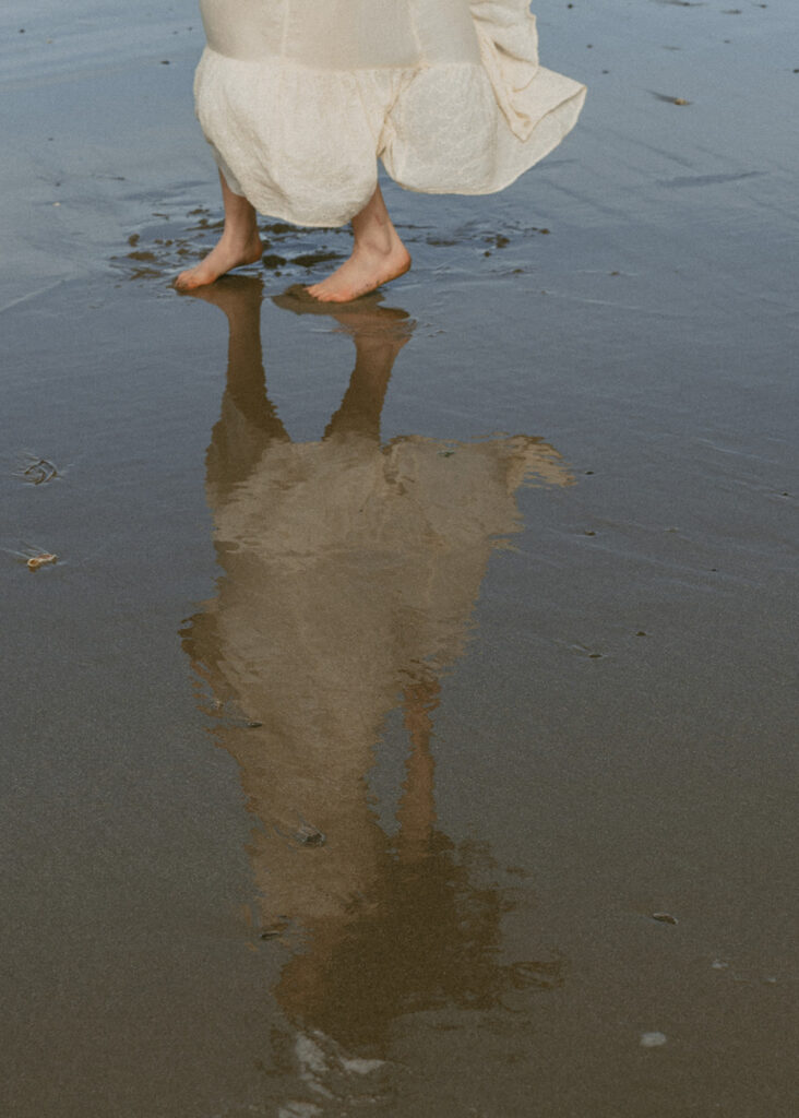 Woman in a flowing white dress standing barefoot on wet sand at the beach, with their reflection visible in a shallow pool of water