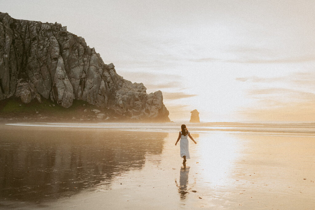 A woman in a white dress walks along a serene beach at sunset for her senior beach photo