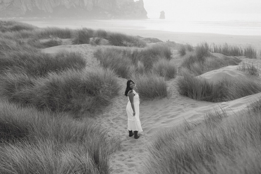 Black and white photo of a woman in a white dress standing in tall grass on a sandy beach, looking back over her shoulder