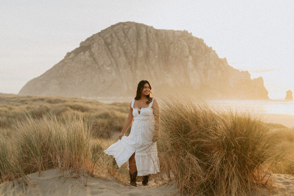 Woman in a white dress standing among tall grassy dunes at the beach during sunset