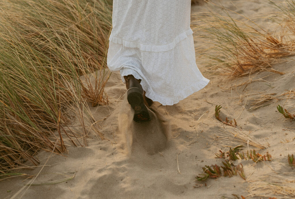 Woman wearing a white dress  with brown shoes that are partially covered with sand walks on a sandy path through tall grass