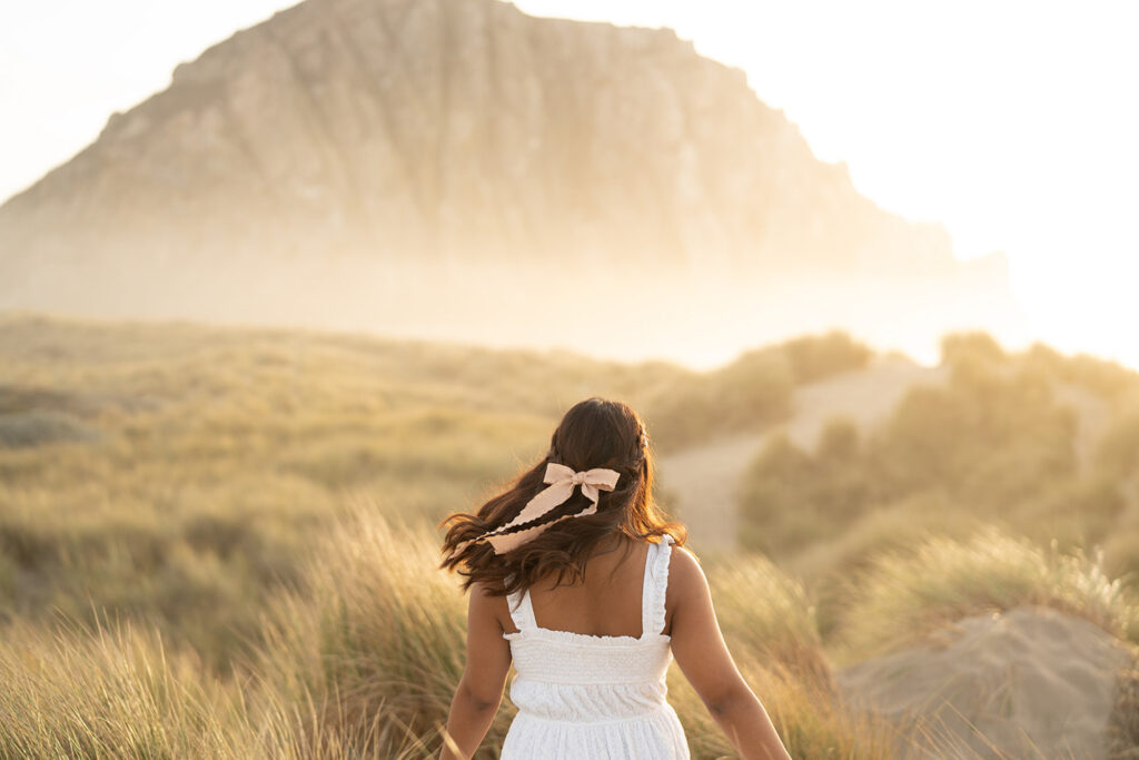 Lady in a white dress with a pink ribbon in their hair walking in a field of tall grass at sunset