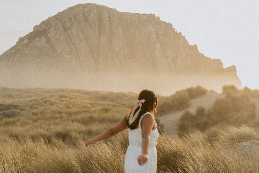 Lady in a white dress with a pink ribbon in their hair stands with arms outstretched in a field of tall grass at sunset