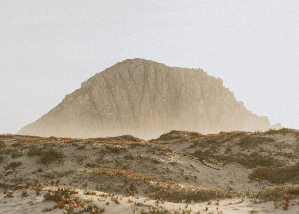 A large, rocky mountain stands in the distance with a hazy, muted sky above it and the foreground features sandy dunes